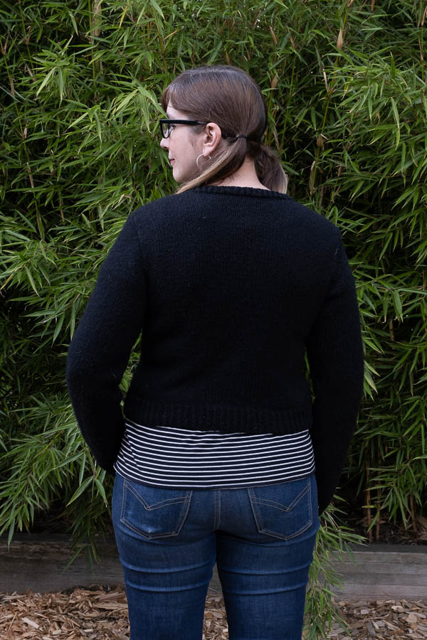 On-figure waist up back shot of a hand knit black cardigan knit with Harrisville Designs Highland yarn and using the CustomFit Inlet Cardigan pattern
